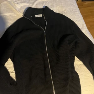 Svart ribbad cardigan jacka  - Snygg svart ribbad cardigan jacka från Filippa K med dragkedja framtill. Perfekt för lager-på-lager och ger en stilren look. Passar bra till både vardag och mer uppklädda tillfällen.
