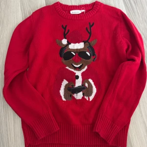 Röd jultröja med renmotiv från KappAhl - Röd stickad tröja från KappAhl med ett broderat renmotiv framtill. Tröjan har rund hals, långa ärmar och ribbade muddar vid ärmslut och nederkant. Tillverkad i 100% bomull – perfekt för julens alla tillfällen. Storlek 134/140