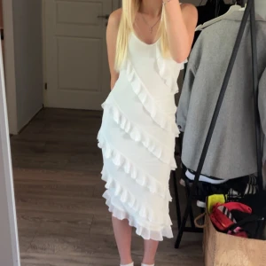 Vit volangklänning med v-ringning - Supersöt vit klänning med v-ringning och volanger i diagonala lager. Klänningen är ärmlös och har en feminin, flowy känsla. Perfekt för dig som vill sticka ut med en romantisk look. Sitter snyggt och har en medellång längd.