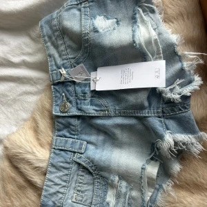 Lågmidjade ljusblå jeansshorts XXS - Snygga lågmidjade jeansshorts från Gina i ljusblå denim. Shortsen har slitna detaljer, fransiga kanter och klassiska fickor både fram och bak. Perfekta för en chill sommarlook med en croppad topp eller hoodie. Stor i storleken!!
