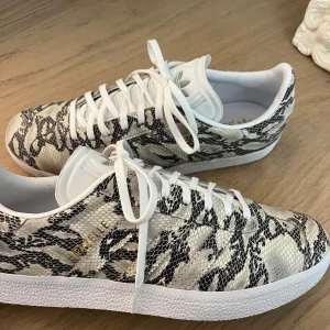 Adidas Gazelle snakeprint sneakers - Snygga Adidas Gazelle sneakers med vit och svart snakeprint. Skorna har klassisk låg siluett, vita snören och detaljer i guld. Skorna är egen designade, oanvända och köpta för 2700kr!!             Säljer pågrund att att det var fel storlek och inte gick att skicka tilbaka när man designat själv.