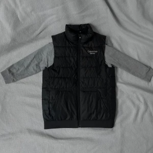 Svart dunväst från Calvin Klein Jeans - Svart quiltad dunväst från Calvin Klein Jeans med hög krage och dragkedja framtill. Västen har två fickor och logga tryckt på bröstet samt längs dragkedjan. Perfekt för lager-på-lager och snygg till både hoodie och t-shirt.