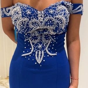 Blå långklänning med pärldetaljer - Magisk blå långklänning med offshoulder och figurnära passform. Klänningen har vackra pärldetaljer i silver över byst, rygg och längs hela kjolen. Utsvängd nederdel ger en elegant siluett, perfekt för bal eller fest. Materialet känns mjukt och följsamt. Passar både S och M! (Har släpp!)