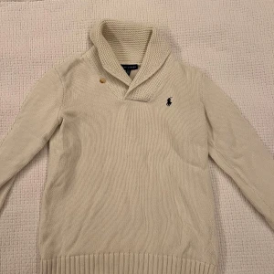 Vit stickad tröja från Ralph Lauren - Säljer en beige stickad tröja från Ralph Lauren med klassisk sjalkrage och en knappdetalj vid halsen. Tröjan har lång ärm, ribbade muddar och den ikoniska broderade loggan på bröstet. Perfekt för dig som gillar stilren och tidlös design.