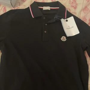 Svart pikétröja från Moncler - Snygg svart pikétröja från Moncler med klassisk krage och knäppning framtill. Tröjan har Moncler-logga på bröstet och diskreta röda och vita detaljer på kragen. Perfekt för en stilren och sportig look. Kan gå ner i pris vid snabb affär 