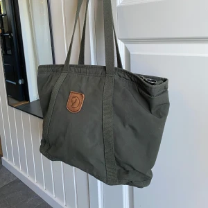 Grön shoppingväska från Fjällräven - Snygg och rymlig grön shoppingväska från Fjällräven med klassisk logga i läder framtill. Väskan har långa handtag och är tillverkad i slitstark bomull/polyester-blandning. Perfekt för att bära med sig allt man behöver under dagen. Går även dra ut axelremmarna på baksidan så att det blir en ryggsäck. 