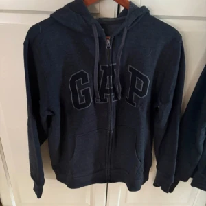 Mörkblå hoodie från GAP med dragkedja - Mörkblå hoodie från GAP med stor logga framtill, dragkedja och huva. Tröjan har känguruficka och är tillverkad i en mjuk blandning av bomull och polyester. Perfekt för chill dagar eller när du vill ha en enkel och snygg look.