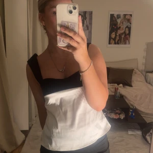 Svartvit korsett-topp med rosett - Säljer en snygg svartvit korsett-topp med breda svarta axelband och knytband i ryggen. Toppen har en cool kontrast mellan svart upptill och vitt nedtill, samt en söt rosett bak. Perfekt för dig som vill sticka ut med en trendig look.