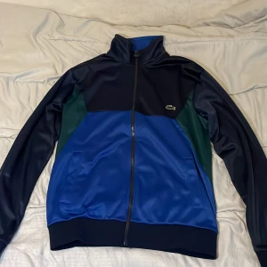 Blå kofta från Lacoste, M - Snygg koftan från Lacoste i blått, mörkblått och grönt. Kofta har hög krage, hel dragkedja och klassisk krokodillogga på bröstet. Regular fit och tillverkad i glansig polyester. Perfekt för dig som gillar sportig och stilren look. Och de är oavvänd
