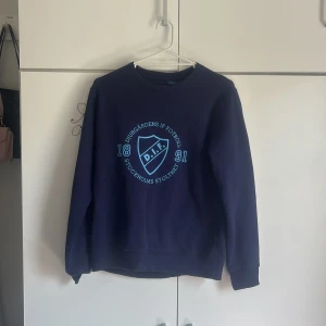 Blå Djurgården sweatshirt M - Mörkblå sweatshirt från Djurgårdens IF med stort ljusblått tryck på bröstet. Klassisk rund halsringning och ribbade muddar vid ärmslut och nederkant. Perfekt för dig som vill visa ditt stöd för DIF. Materialet är mjukt och skönt bomullsmix.