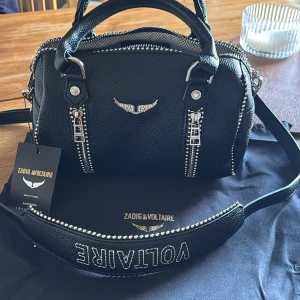 Svart handväska Zadig & Voltaire skinn - Cool svart handväska från Zadig & Voltaire i grymt snyggt skinn. Väskan har silvriga detaljer, dubbla dragkedjor framtill och ikonisk vinge i metall. Perfekt storlek för det viktigaste och med ett stadigt handtag. Kommer med original dustbag och tag.