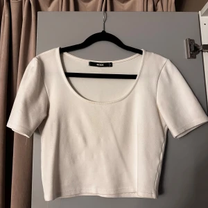 Vit croppad topp från Bik Bok - Enkel och stilren vit croppad topp från Bik Bok med rundad, djup urringning och korta ärmar. T-shirten är i mjuk bomull och passar perfekt till jeans eller kjol för en clean look.
