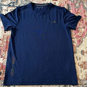 Blå Under Armour träningst-shirt M - Snygg marinblå t-shirt från Under Armour i tekniskt material med diskret struktur. Klassisk passform med korta ärmar och rund hals. Perfekt för träning, med logga på bröstet och tryck längs sidan. Lätt och snabbtorkande för aktiva dagar.