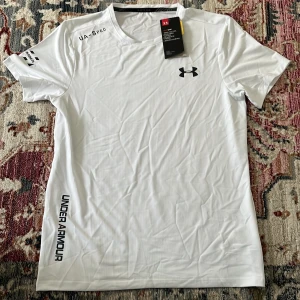Vit Under Armour träningst-shirt - Snygg vit t-shirt från Under Armour med svart logga på bröstet och tryck på ärm och sida. Tillverkad i lätt och ventilerande polyester, perfekt för träning. Klassisk passform med korta ärmar och rund hals. Stilren och sportig vibe.