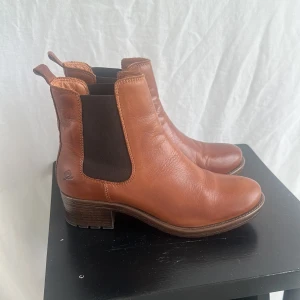 Bruna chelsea boots från Rosa Negra - Snygga bruna chelsea boots i skinn från Rosa Negra. Skorna har elastiska svarta paneler på sidorna och en låg klack i trälook. Perfekta för dig som gillar klassisk stil med en modern touch.nn