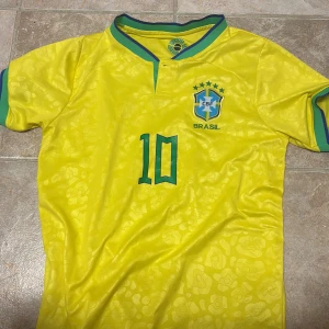 Brasilien Neymar Jr 10 matchtröja - Gul Brasilien fotbollströja från Nike med gröna detaljer, Neymar Jr och nummer 10 på ryggen. Tröjan har korta ärmar, CBF-emblem på bröstet och subtilt mönster i tyget. Perfekt för dig som älskar fotboll och vill sticka ut på läktaren eller planen.