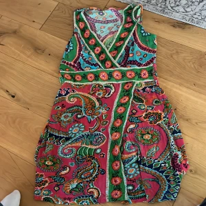 Färgglad omlottklänning i paisleymönster - Säljer en superfin kort omlottklänning med färgstarkt paisleymönster i grönt, rosa, blått och orange. Klänningen har v-ringning, markerad midja och är ärmlös. Perfekt för dig som vill sticka ut med en unik och bohemisk stil.