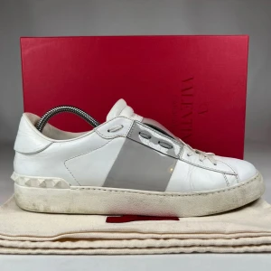 Valentino Open - Snygga Valentino Garavani Open sneakers i vitt skinn med grått lackat band över sidan. Klassisk låg modell med snörning och ikoniska nitar på hälen. Perfekta för dig som gillar lyxiga detaljer och stilren design. Pris kan diskuteras vid snabb affär.