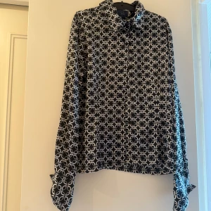 Marinblå mönstrad skjorta från RAG - Snygg marinblå skjorta från RAG i storlek M med vitt grafiskt mönster. Skjortan är tillverkad i glansig polyester och har klassisk krage, långa ärmar och knäppning framtill. Perfekt för dig som gillar att sticka ut med coola prints.