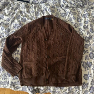 Brun kabelstickad kofta Polo Ralph Lauren - Säljer en brun kabelstickad kofta från Polo Ralph Lauren. Koftan har v-ringning, knappar framtill, två fickor och långa ärmar. Klassisk och snygg modell i mjukt stickat material, perfekt för lager-på-lager-stil. Köpt på Ralph Laurens huvudbutik i London för 3 veckor sedan (Ord pris 3700kr). Endast använd en gång🙃
