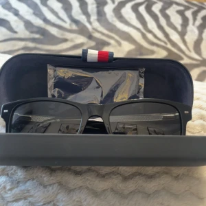 Svarta solglasögon Tommy Hilfiger - Stilrena svarta solglasögon från Tommy Hilfiger med klassisk rektangulär form och diskreta silvriga detaljer på skalmarna. Säljs med originalfodral och putsduk, perfekt för dig som gillar en clean och tidlös look.