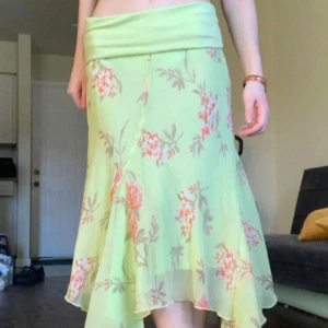 Grön blommig långkjol med volang - Superfin ljusgrön långkjol med blommigt mönster i rosa och orange toner. Kjolen har volangig nederkant och är gjord i ett lätt, flowy material som känns perfekt för vår och sommar. Midjan är bred och stretchig för extra bekvämlighet.