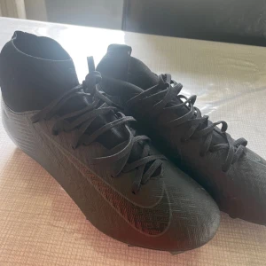 Svarta Nike fotbollsskor med snörning - Snygga helsvarta Nike fotbollsskor med högre strumpliknande skaft och snörning. Skorna har en mönstrad ovandel i syntetmaterial och dobbar för bra grepp på planen. Perfekta för dig som vill ha en clean look på matchen.