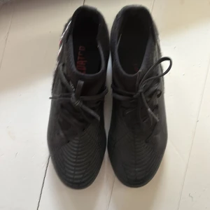 Adidas Predator svarta fotbollsskor - Säljer ett par svarta Adidas Predator fotbollsskor med klassiska tre ränder i vitt och rött på sidan. Skorna har snörning, låg profil och mönstrad ovandel i syntetmaterial. Sulan har blå dobbar för bra grepp på planen. Perfekta för dig som vill sticka ut på matchen.