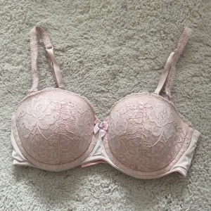 Rosa spetsblus från Hunkemöller - Supersöt ljusrosa blus från Hunkemöller med blommig spets, vadderade kupor och fina rosett-detaljer. Axelbanden är dekorerade och blusen har en liten metallring framtill. Perfekt för dig som gillar romantisk och feminin stil. Man kan även knäppa av banden på bh