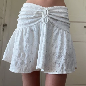 Vit kortkjol med snörning - Superfin vit kortkjol med snygg snörning framtill och rynkad midja. Kjolen har en lätt volang och är gjord i ett tunt, strukturerat bomullstyg som känns luftigt. Perfekt för varma dagar och ger en fräsch sommarkänsla. De har inbyggda shorts!