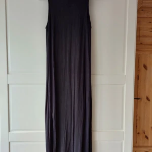 Svart långklänning från Lindex - En stilren svart långklänning från Lindex med ärmlös design och hög krage. Klänningen har en rak och enkel siluett i mjukt material, perfekt för en clean och modern look. Passar dig som gillar minimalistisk stil och vill ha något bekvämt men ändå snyggt. Den har slitsar i båda sidorna. 