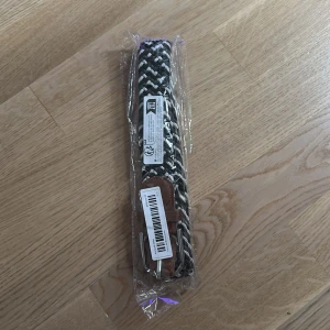 Svartvitt flätat bälte med skinn - Snyggt flätat bälte i svart och vitt med bruna skinn-detaljer och silverspänne. Perfekt accessoar för att lyfta din outfit med en cool och modern touch. Bältet har ett randigt mönster och är flexibelt tack vare den stickade designen.