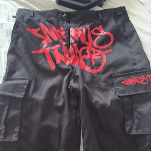 Svarta cargopants med röd graffiti - Svarta cargopants med stora fickor på sidorna och fet röd graffiti-text framtill. Byxorna har en loose fit och cool streetkänsla, perfekta för dig som gillar att sticka ut. Materialet känns slitstarkt och trycket är riktigt iögonfallande.