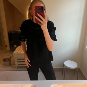 Svart blus med volang från & Other Stories - Supersöt svart blus från & Other Stories med broderade detaljer, volang vid hals och ärmar samt kort ärm. Blusen har en spetsinslag och rund halsringning.