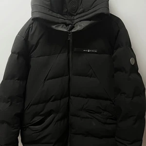 Svart pufferjacka från Sail Racing - Passar storlek M-L, Svart pufferjacka från Sail Racing med huva och dragkedja framtill. Jackan har två snedställda fickor med dragkedja och diskret logga på bröstet. Perfekt för kalla dagar med sin vadderade design och stilrena look.