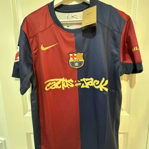 FC Barcelona x Travis scott Nike tröja - Säljer en FC Barcelona x Cactus Jack matchtröja från Nike. Tröjan har klassisk halv röd och halv marinblå design med gul Nike-logga och Cactus Jack-tryck på bröstet. Broderat Barca-märke och patch på ärmen. Tillverkad i lätt, ventilerande material.