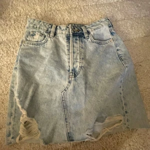 Ljusblå jeanskjol med slitningar - Trendig ljusblå jeanskjol med slitna detaljer och fransar längst ner. Klassisk femficksmodell med bälteshällor och dragkedja framtill. Nästan inte använd! 
