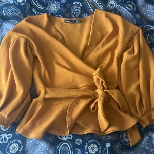 Omlottopp i senapsgult från Boohoo - Superfin omlottopp i senapsgult från Boohoo med puffiga långärmar och knyt i midjan. Toppens v-ringning och volangkant ger en trendig look. Perfekt för dig som vill sticka ut med färg och snygg passform.