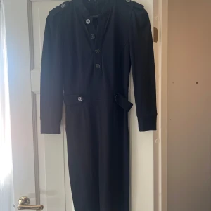 Svart mellanlång från Stefanel - Stilren svart långklänning från Stefanel med markerade axlar, långa ärmar och snygga knapprader framtill. Klänningen har dekorativa knappar i midjan och axelpartiet samt dragkedja i ryggen. Perfekt för dig som gillar en klassisk och elegant look.