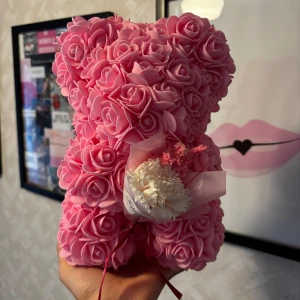 Rosbjörn - Bilderna visar en söt nallebjörn formad av rosa konstgjorda rosor, dekorerad med en liten bukett blommor. Den är perfekt som present eller dekoration.