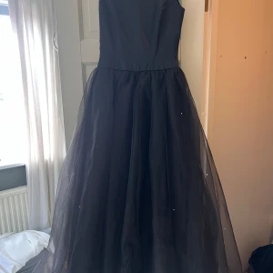 Svart långklänning från Vila - Supersnygg svart långklänning från Vila med ärmlös överdel och voluminös kjol i tyll. Klänningen har diskreta små kristaller på kjolen och dragkedja i ryggen. Perfekt för bal eller festliga tillfällen. Materialet är polyester och nylon.