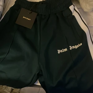 Palm Angels svarta shorts med vit stripe - Palm Angels shorts i svart med vit sidoremsa och broderad logga framtill. Elastisk midja med snörning för skön passform. Sportig och clean look, perfekt för dig som gillar streetwear och vill sticka ut med en ikonisk detalj.