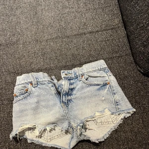 Ljusblå jeansshorts från Gina Tricot - Snygga ljusblå jeansshorts från Gina Tricot med fransiga kanter och slitna detaljer. Klassisk femficksmodell med bälteshällor och knappgylf. Perfekta för varma dagar när du vill ha en avslappnad och trendig look.