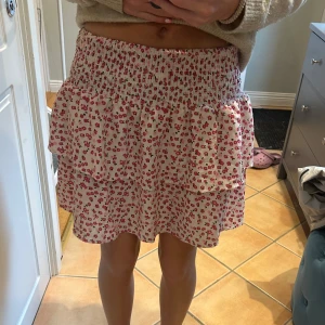 Blommig volangkjol med smock - Supersöt kort kjol med volanger och smock i midjan. Vit botten med små rosa blommor över hela kjolen. 