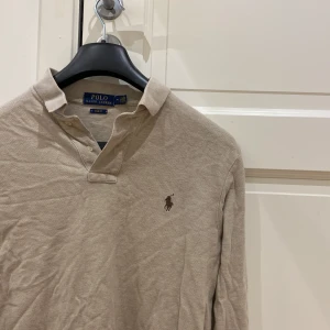 Beige långärmad pikétröja Polo Ralph Lauren - Klassisk beige långärmad pikétröja från Polo Ralph Lauren med brunt broderat logotyp på bröstet. Tröjan har krage, knappar vid halsen och är i bomull. Slim fit-modell som passar perfekt till jeans eller chinos.