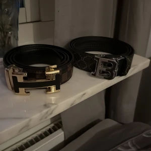 Två designerbälten läder - Säljer två snygga bälten med stora metallspännen, ett med guldigt H och ett med silvrigt B. H-bältet har brunt och svart rutigt mönster, B-bältet är mörkgrått med diskret mönster och B-detaljer. Båda är i skinn och har exklusiv vibe, perfekta för att lyfta din outfit.