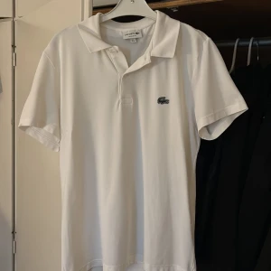 Vit pikétröja från Lacoste - Klassisk vit pikétröja från Lacoste med ikoniska krokodilloggan på bröstet. Tröjan har korta ärmar, krage och knäppning framtill. Tillverkad i bomull med regular fit som ger en clean och stilren look.