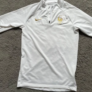Vit långärmad Nike Frankrike-tröja - Snygg vit långärmad fotbollströja från Nike med Frankrikes landslagsemblem och två stjärnor broderade i guld på bröstet. Tröjan har dragkedja vid halsen och är tillverkad i ett mjukt, sportigt material som andas. Perfekt för dig som gillar stilren design och fotboll.