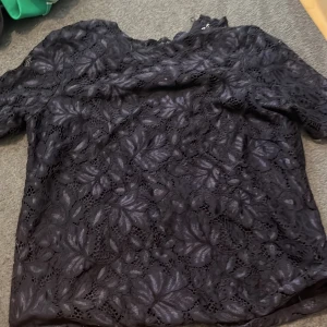 Svart spetsblus med blommönster S - Säljer en svart blus i spets med blommigt mönster. Toppen har korta ärmar och är fodrad undertill, vilket ger en snygg kontrast till det genombrutna spetsmaterialet. Perfekt för dig som gillar detaljer och en lite elegantare stil.