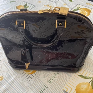 Louis Vuitton Vernis Alma BB Bag - Snygg svart handväska från Louis Vuitton i glansigt skinn med klassiskt LV-monogram över hela väskan. Väskan har guldfärgade metalldetaljer, dubbla handtag och dragkedja med lås. Perfekt för dig som vill ha en lyxig och tidlös accessoar.
